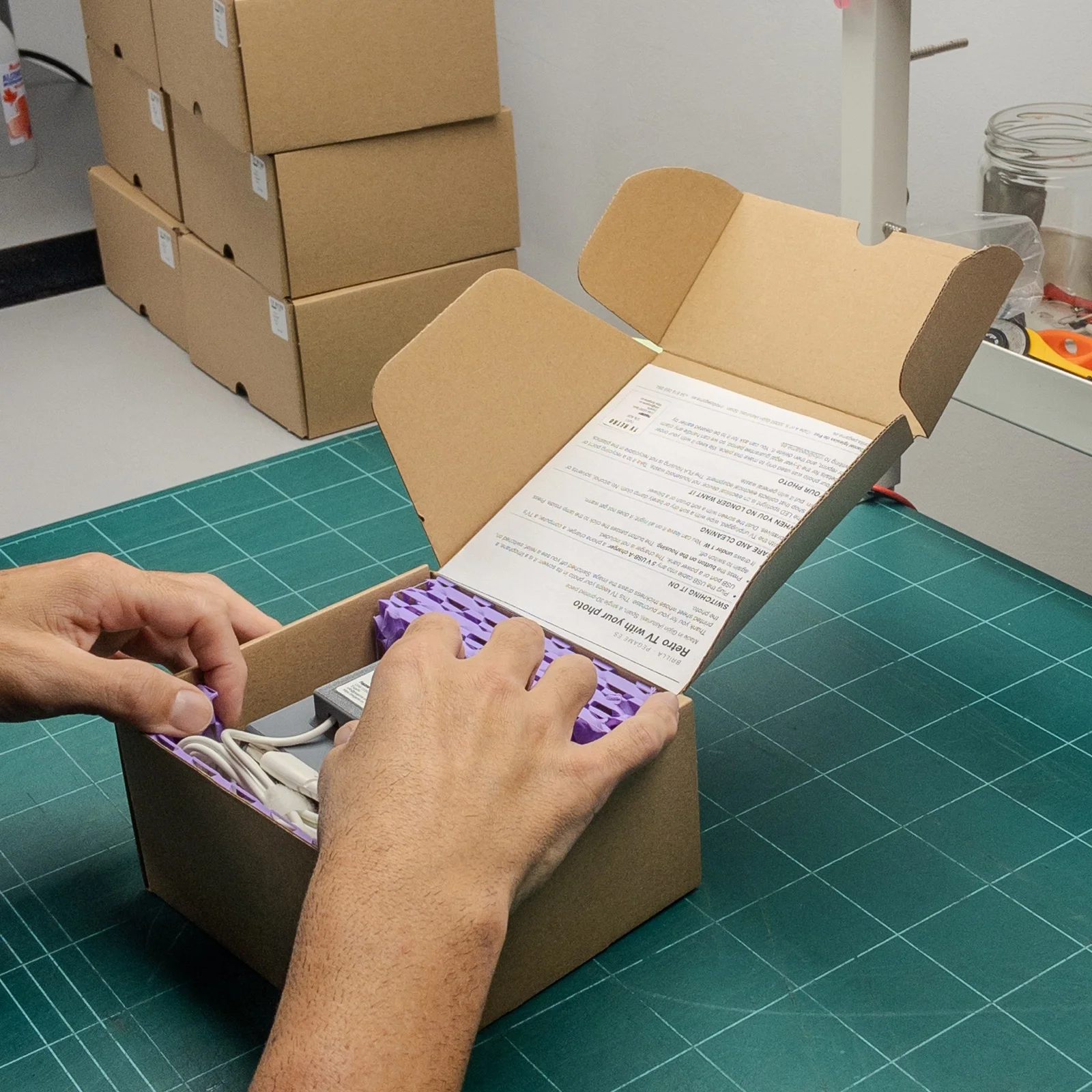 Manos embalando una pieza impresa en 3D en una caja de cartón con protector morado, con más cajas apiladas detrás