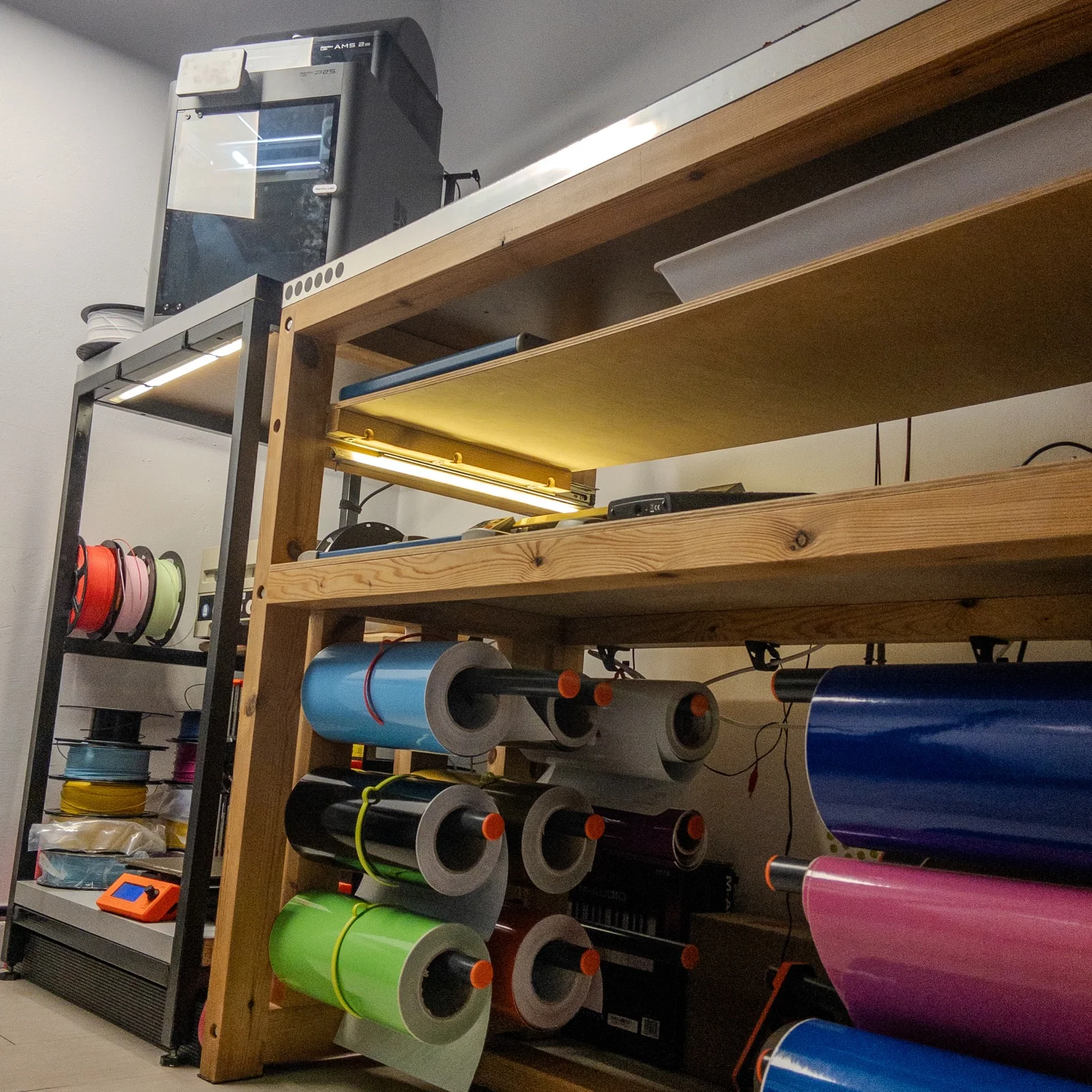 Taller visto desde abajo: impresoras 3D y bobinas de filamento junto a una estantería de madera llena de rollos de vinilo de colores