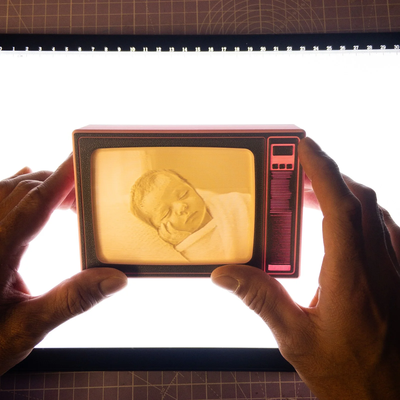 Manos sujetando a contraluz una pieza impresa en 3D con forma de televisor retro que muestra la foto de un bebé dormido