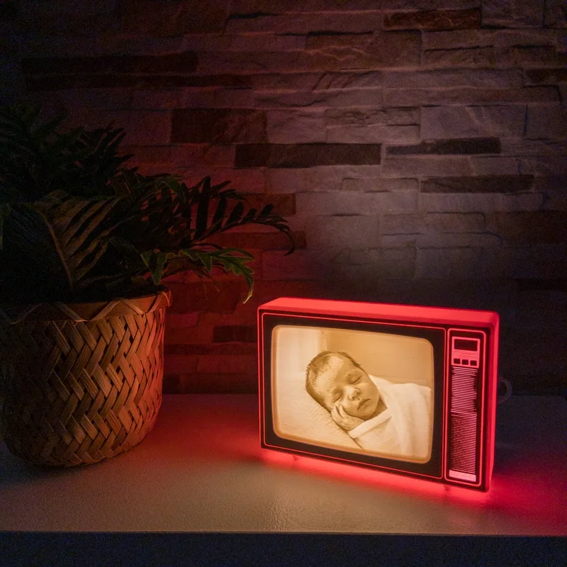 TV retro de Brilla roja encendida en una estantería junto a una planta, con la foto de un bebé dormido iluminada en luz cálida