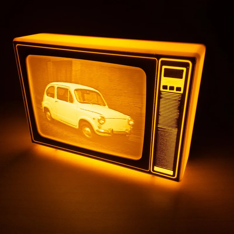 TV retro amarilla de Brilla encendida en la oscuridad; la luz cálida tiñe la carcasa de naranja y dibuja en la pantalla abombada la foto de un Seat 600 aparcado junto a una pared