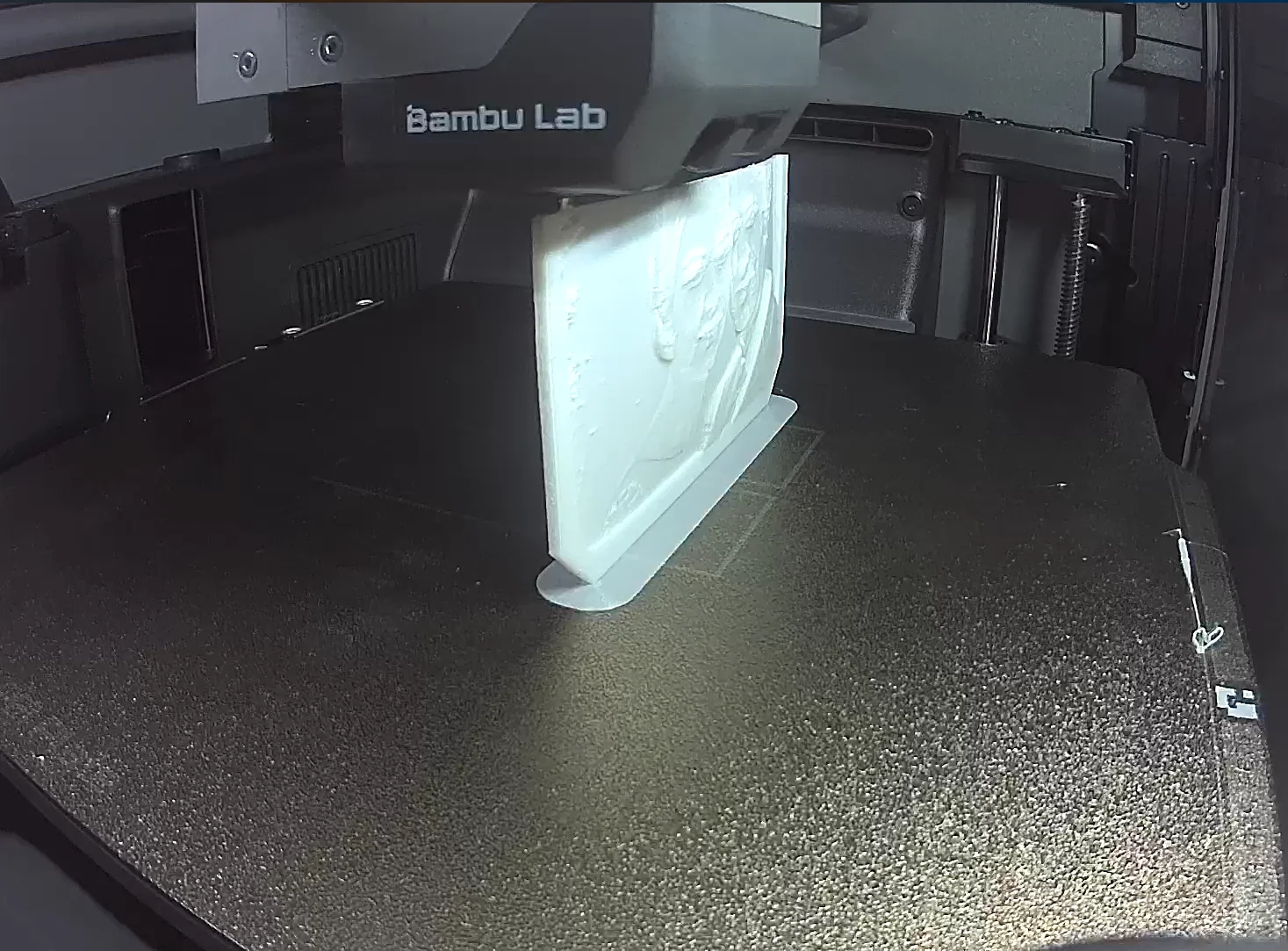Inside a 3D printer in the workshop, a half-printed white lithophane stands upright on the print bed; the faces of a photo can already be made out in its relief