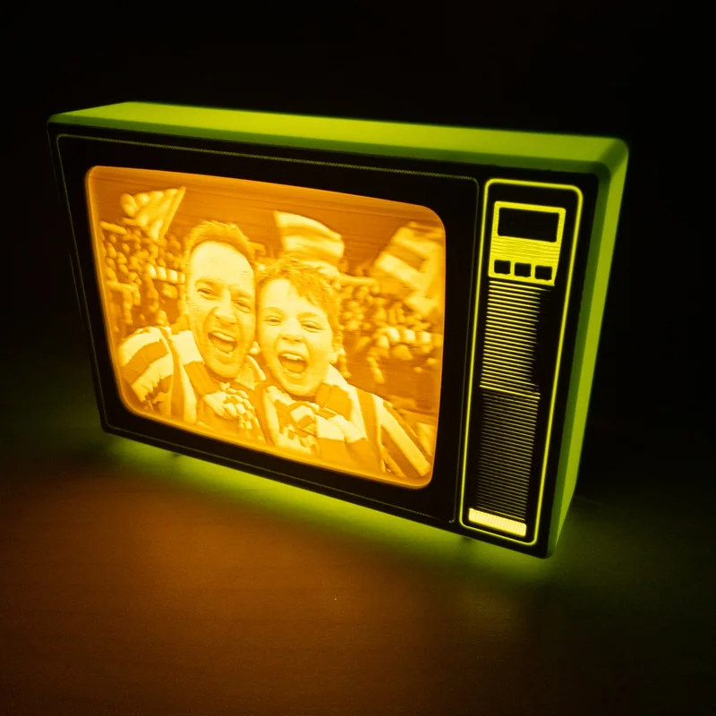 Green retro TV lit up in the dark with a lithophane of a father and son shouting with joy in the stands of a stadium, wearing striped scarves, in amber tones